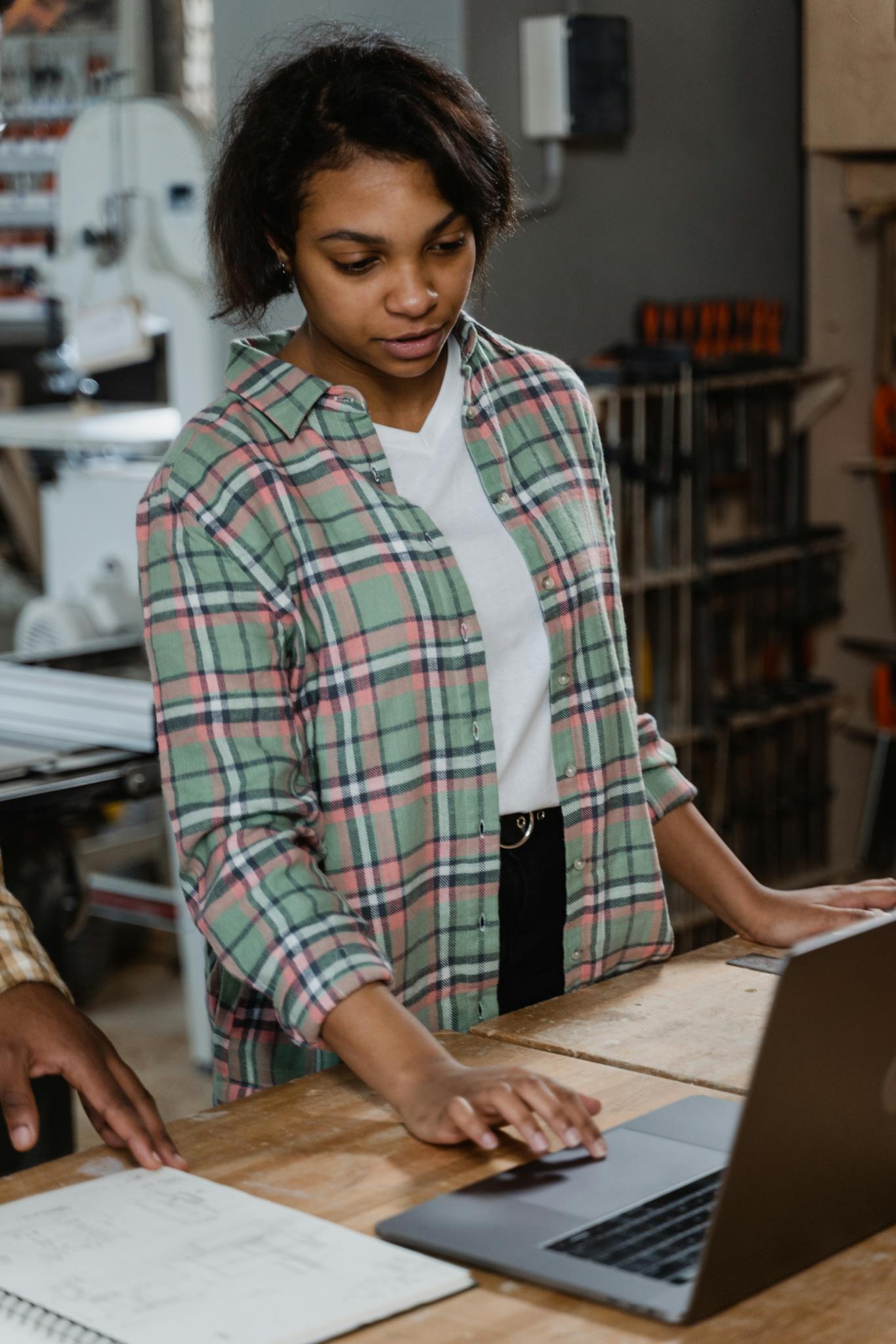 A young woman in a workshop using a laptop to manage woodworking tasks.
